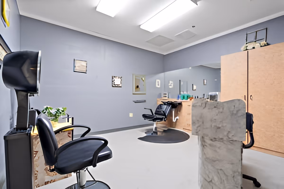 Interior of a salon room with two black salon chairs, a hair dryer, a large mirror on the wall, a wooden cabinet, and various hair care products on the counter. The walls are painted light blue and the floor is white.