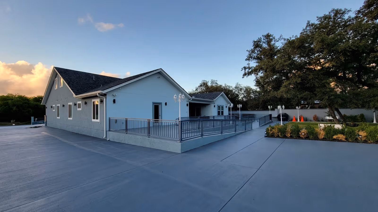 Exterior view of a single-story assisted living facility building with a gray roof and light-colored walls. The building has multiple windows and a ramp with railings leading to the entrance. There are trees and shrubs around the paved area, and the sky is clear with some clouds at sunset.