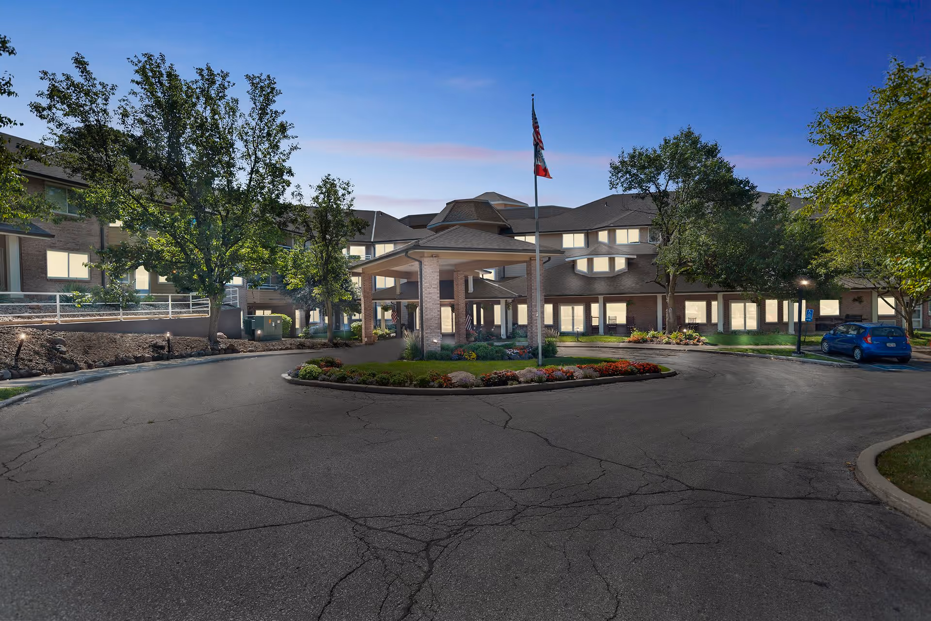 Exterior view of Independence Village of Des Moines at dusk, showing the main entrance with a covered drop-off area, a circular driveway with landscaped center island, trees, and a flagpole with flags. The building is multi-story with many windows illuminated from inside.