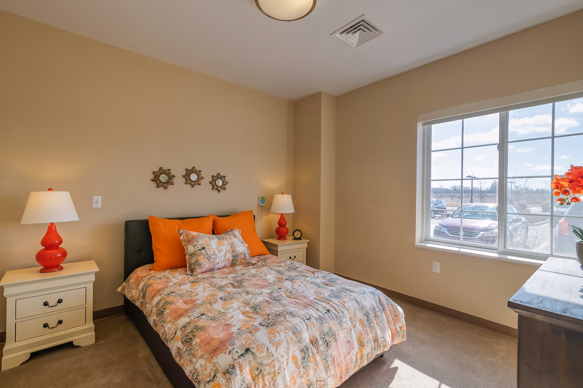 A cozy bedroom with a bed featuring a floral and orange patterned comforter and orange pillows. There are two white nightstands on either side of the bed, each with a red lamp. Three decorative sun-shaped mirrors hang on the beige wall above the bed. A large window on the right side lets in natural light and shows a view of parked cars outside. A wooden dresser with a potted plant is partially visible on the right.