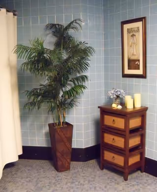 Corner of a tiled interior room with a tall potted palm, a small wooden three-drawer cabinet topped with candles and a framed wall print.