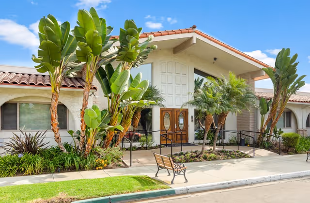 Exterior view of Brittany House Residential Memory Care facility featuring a beige stucco building with a red tile roof, large tropical plants and palm trees near the entrance, a wooden double door with glass panels, a bench on the sidewalk, and a clear blue sky.
