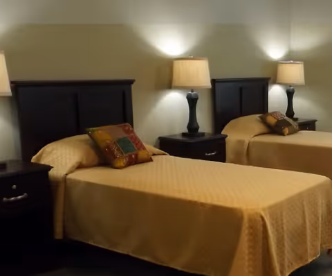 A bedroom with two single beds, each with a dark wooden headboard, beige bedspreads, and a decorative pillow. Between the beds are two dark wooden nightstands, each with a lamp emitting soft light against a beige wall.