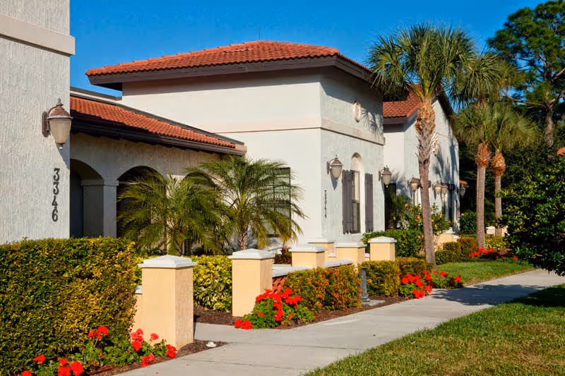 Exterior view of a senior living facility building with white walls and red tile roof, surrounded by palm trees, bushes, and red flowers along a sidewalk under a clear blue sky.