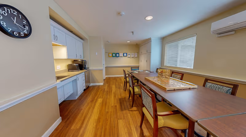 A well-lit room with a long wooden table surrounded by chairs, a large window with blinds on the right wall, and a kitchenette area with white cabinets, a toaster, and a coffee maker on the left. A clock is mounted on the left wall, and framed artwork is displayed on the far wall.