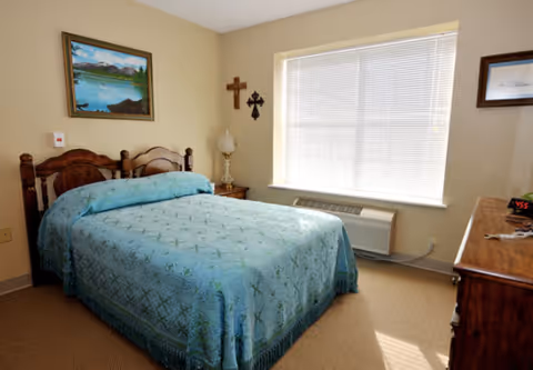 A bedroom with a wooden double bed covered with a blue patterned bedspread. There is a nightstand with a lamp and two crosses hanging on the wall above it. A large window with blinds lets in natural light. A wooden dresser with a digital clock and keys is on the right side of the room. The walls are light-colored and there is a framed painting above the bed.