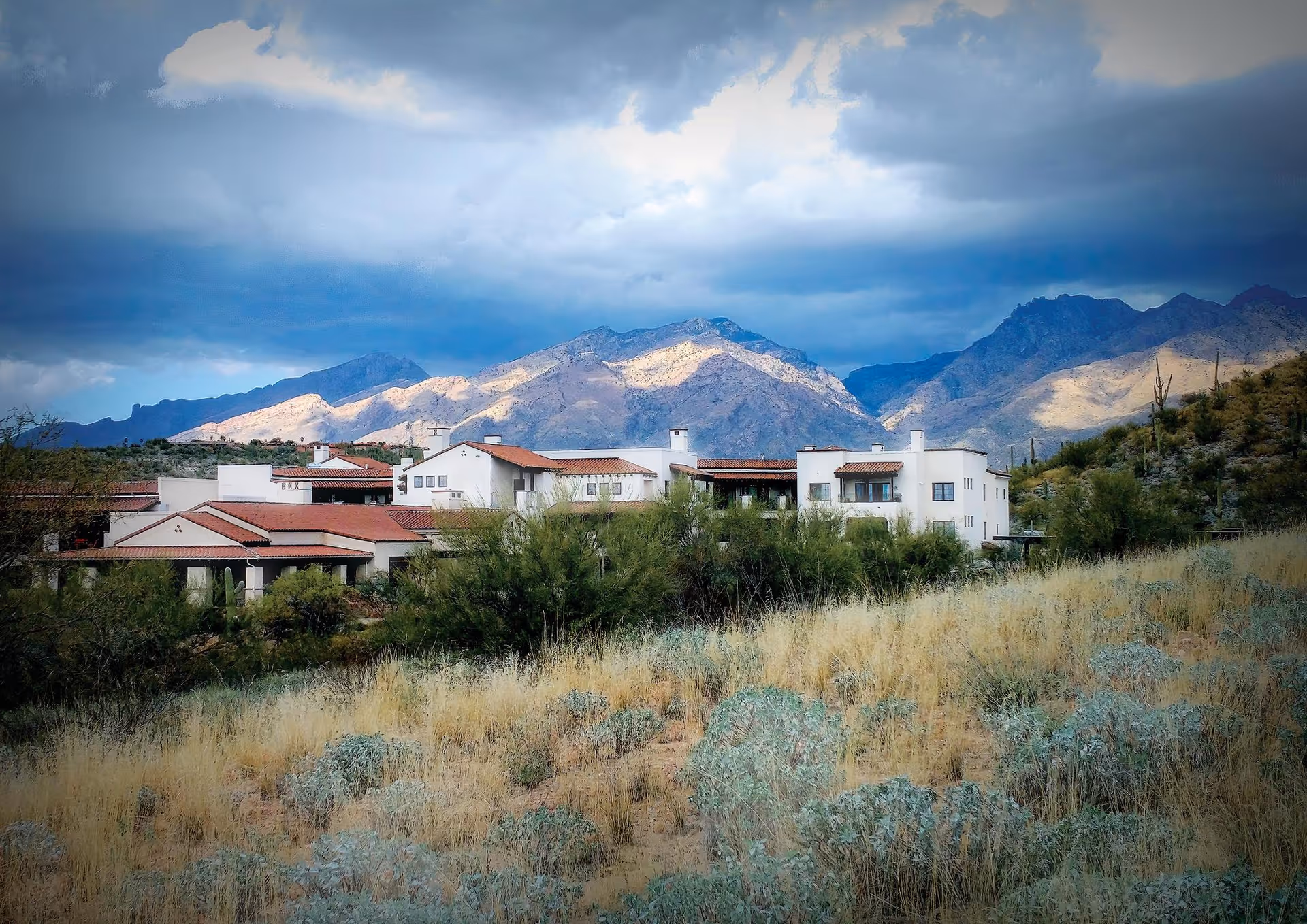 White hacienda-style building with red tile roofs nestled in desert scrub with mountains and a cloudy sky in the background.
