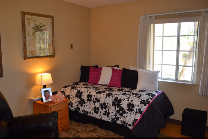 A cozy bedroom with a single bed covered in a black and white floral bedspread with pink and white pillows. There is a wooden nightstand with a lamp, a framed photo, and some flowers. A window with light curtains lets in natural light, and a framed picture of a palm tree hangs on the wall. A black leather chair is partially visible in the foreground.