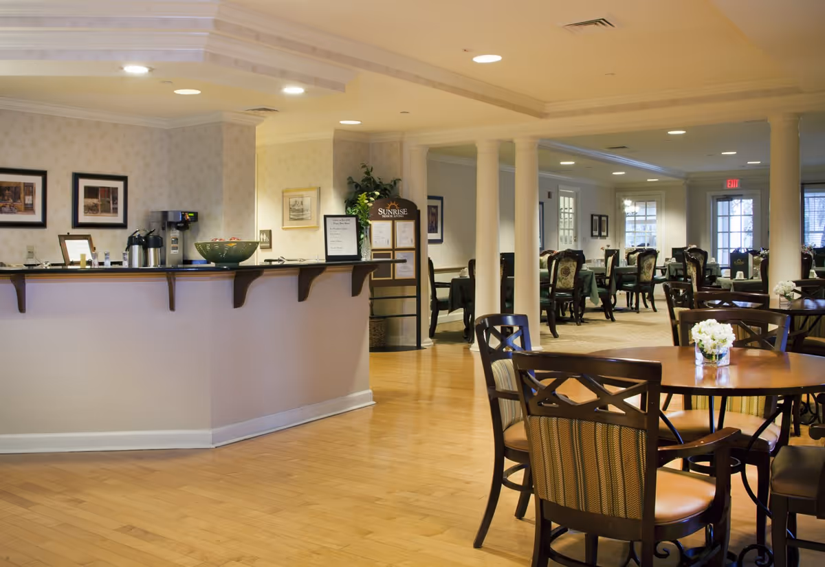 Interior view of a senior living facility dining area with wooden tables and chairs, a counter with coffee dispensers and a fruit bowl, decorative columns, framed pictures on the walls, and soft lighting.