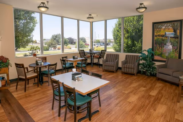 Bright dining and lounge area with tables, chairs, armchairs, and large windows overlooking landscaped grounds.