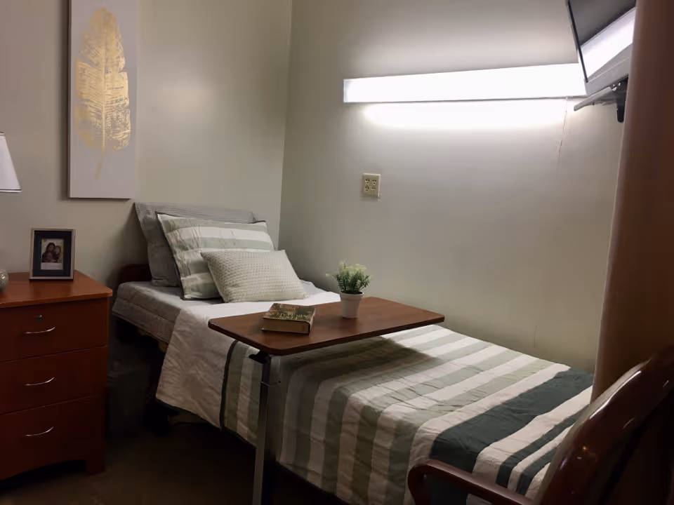 Small single bedroom with a striped bed, overbed table holding a plant and book, a nightstand with a lamp and framed photo, and a wall-mounted light.