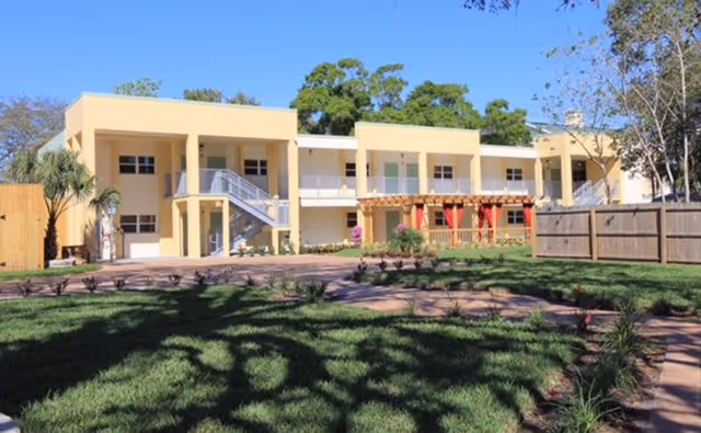 Exterior view of a two-story yellow senior living facility building with balconies and outdoor staircases, surrounded by a green lawn, trees, and a wooden fence under a clear blue sky.