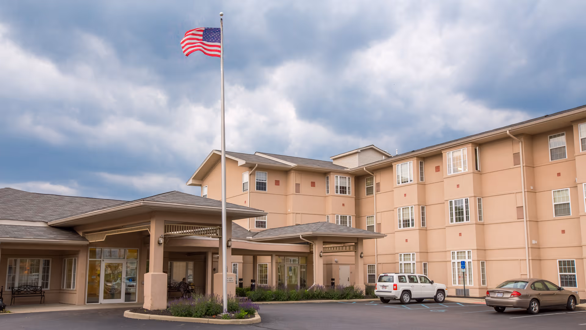 Exterior view of a multi-story senior living facility building with beige walls and multiple windows. There is a covered entrance with a flagpole flying the American flag in front. Several cars are parked in the parking lot near the entrance under a cloudy sky.