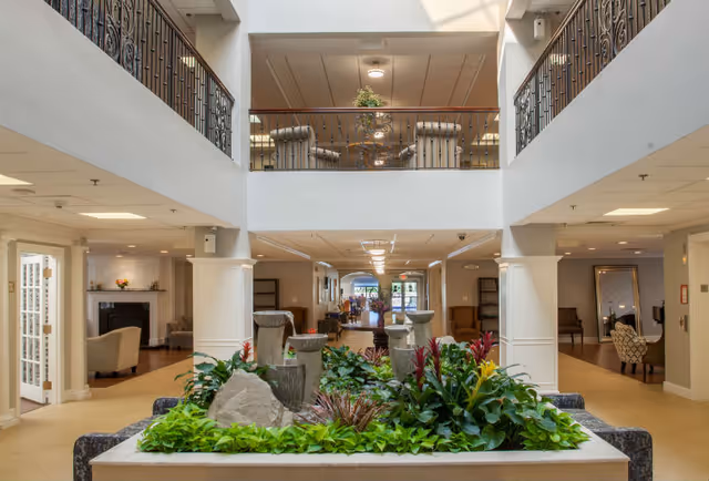 Interior view of a senior living facility lobby with a central indoor garden featuring green plants and flowers. The space has a high ceiling with a second-floor balcony overlooking the area. Comfortable seating areas with armchairs are visible on both sides, and the hallway extends toward the back with more seating and decorative elements.