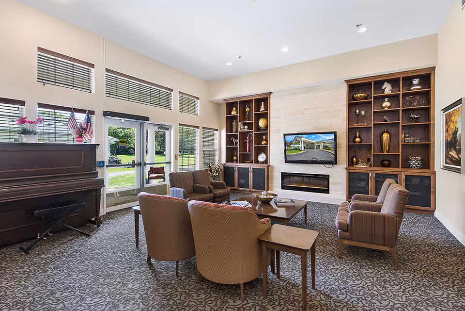A bright and spacious living room in Deerfield Village Assisted Living featuring a patterned carpet, a piano with flowers and small American flags on top, several upholstered armchairs arranged around a wooden coffee table, built-in wooden shelves with decorative items, a wall-mounted flat screen TV above a modern fireplace, and large windows and glass doors letting in natural light with a view of greenery outside.