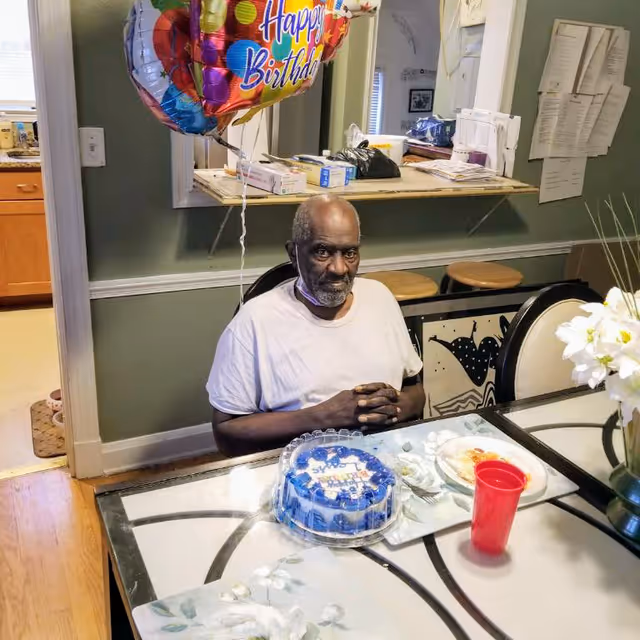 An elderly man sitting at a dining table with a birthday cake in front of him. There are colorful 'Happy Birthday' balloons attached to the chair behind him. The table also has a red plastic cup, a plate with some food, and a vase with white flowers. The background shows a kitchen area and some papers pinned on the wall.