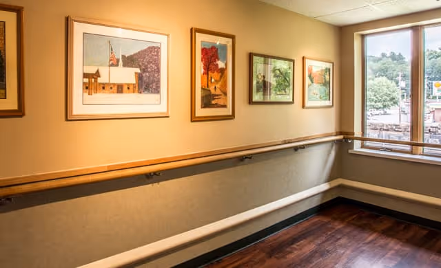 A hallway in a senior living facility with wooden handrails along the walls, several framed paintings hanging on the wall, and a large window showing trees and buildings outside.