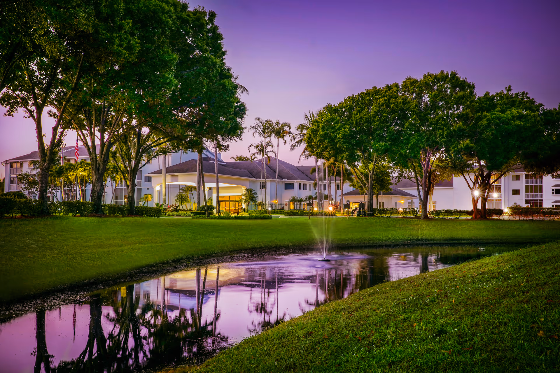 A landscaped lawn and pond with a fountain in front of a low-rise residential building framed by trees at dusk.