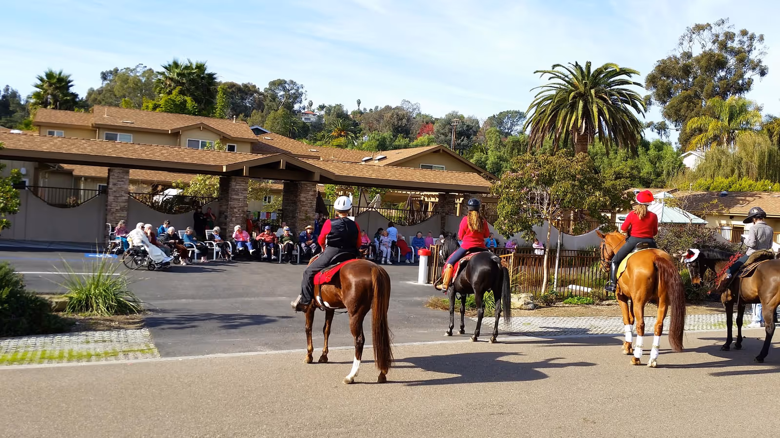 Several horseback riders approach a group of seated senior residents gathered outside the front of a senior living facility.