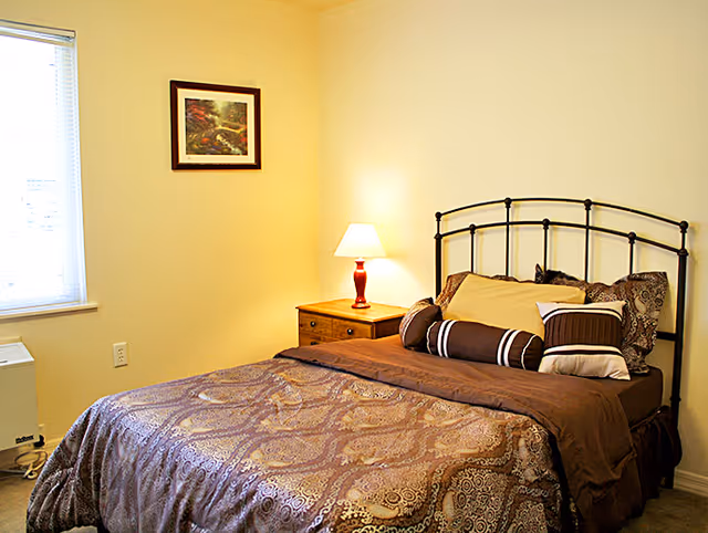 A cozy bedroom with a bed featuring a metal headboard, multiple pillows, and a patterned brown bedspread. There is a wooden nightstand with a red lamp on it, a framed picture on the wall, and a window with blinds partially open letting in natural light.