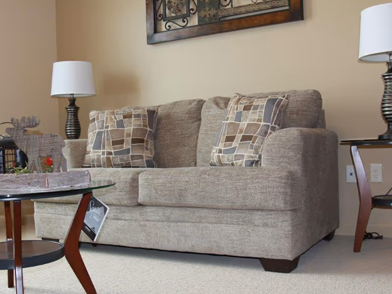 Beige upholstered loveseat with patterned throw pillows, table lamps, wall art, and a glass coffee table in a living room.