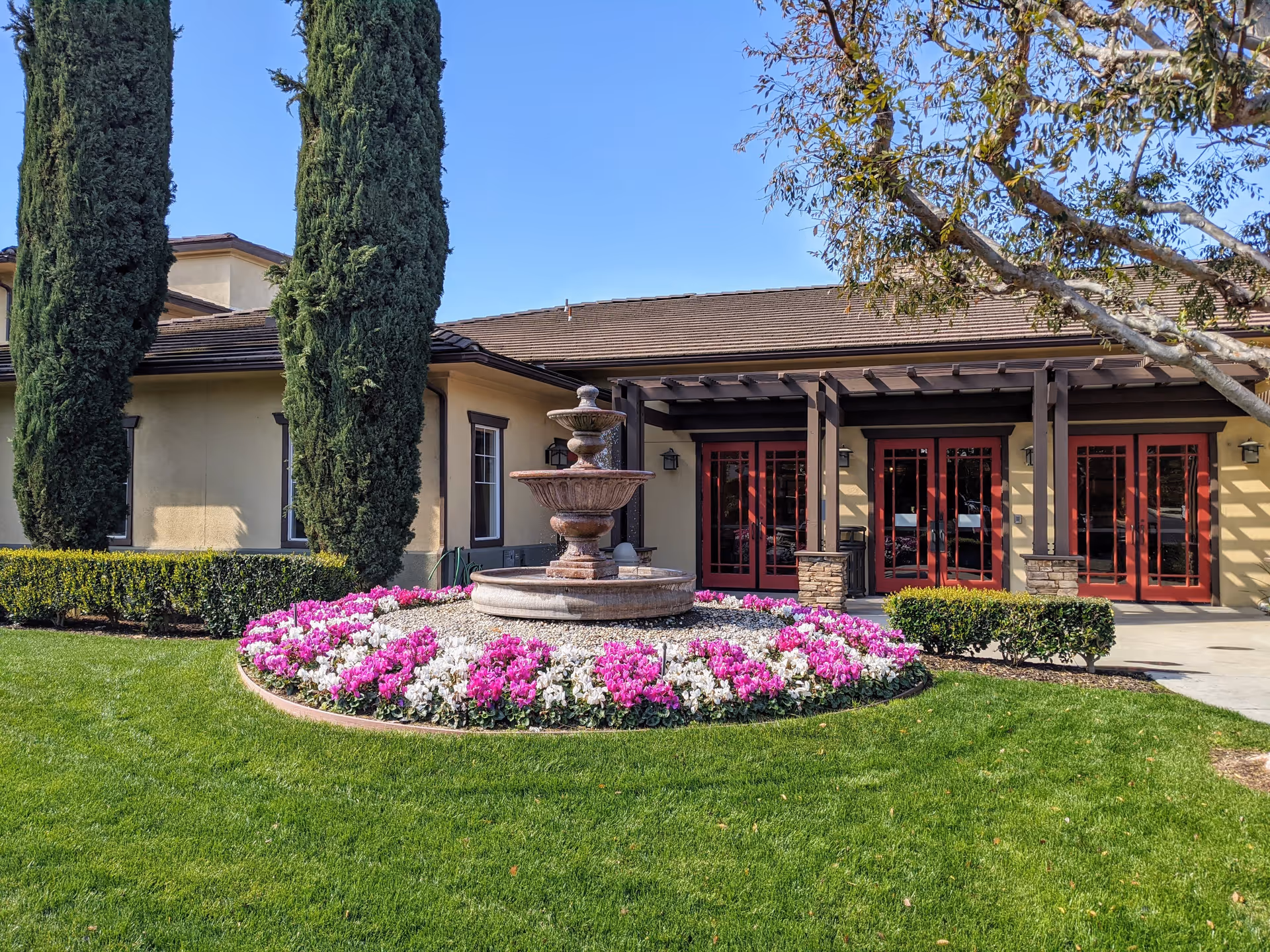 Outdoor view of Woodbridge Terrace facility showing a well-maintained garden with green grass, a circular flower bed with pink and white flowers surrounding a tiered stone fountain, tall cypress trees, and a building with beige walls, red-framed glass doors, and a pergola.