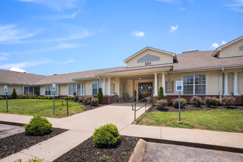 Front exterior view of a single-story senior living facility building with beige siding and brick accents, a covered entrance with columns, rocking chairs on the porch, and a clear blue sky above. There are reserved handicap parking signs and landscaped bushes along the walkway leading to the entrance.