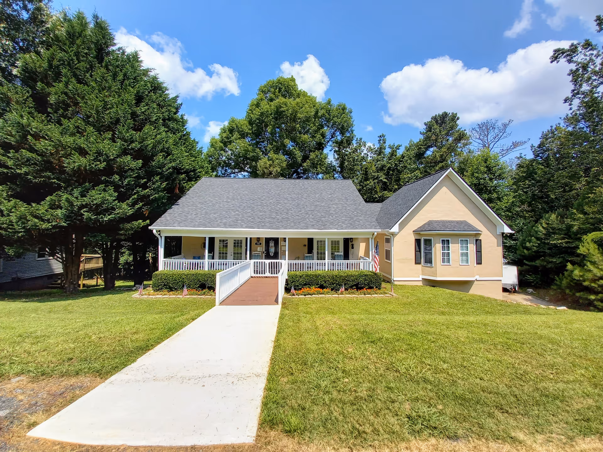 Single-story pale yellow building with a covered front porch, wheelchair ramp, American flag, and a concrete walkway across a green lawn.