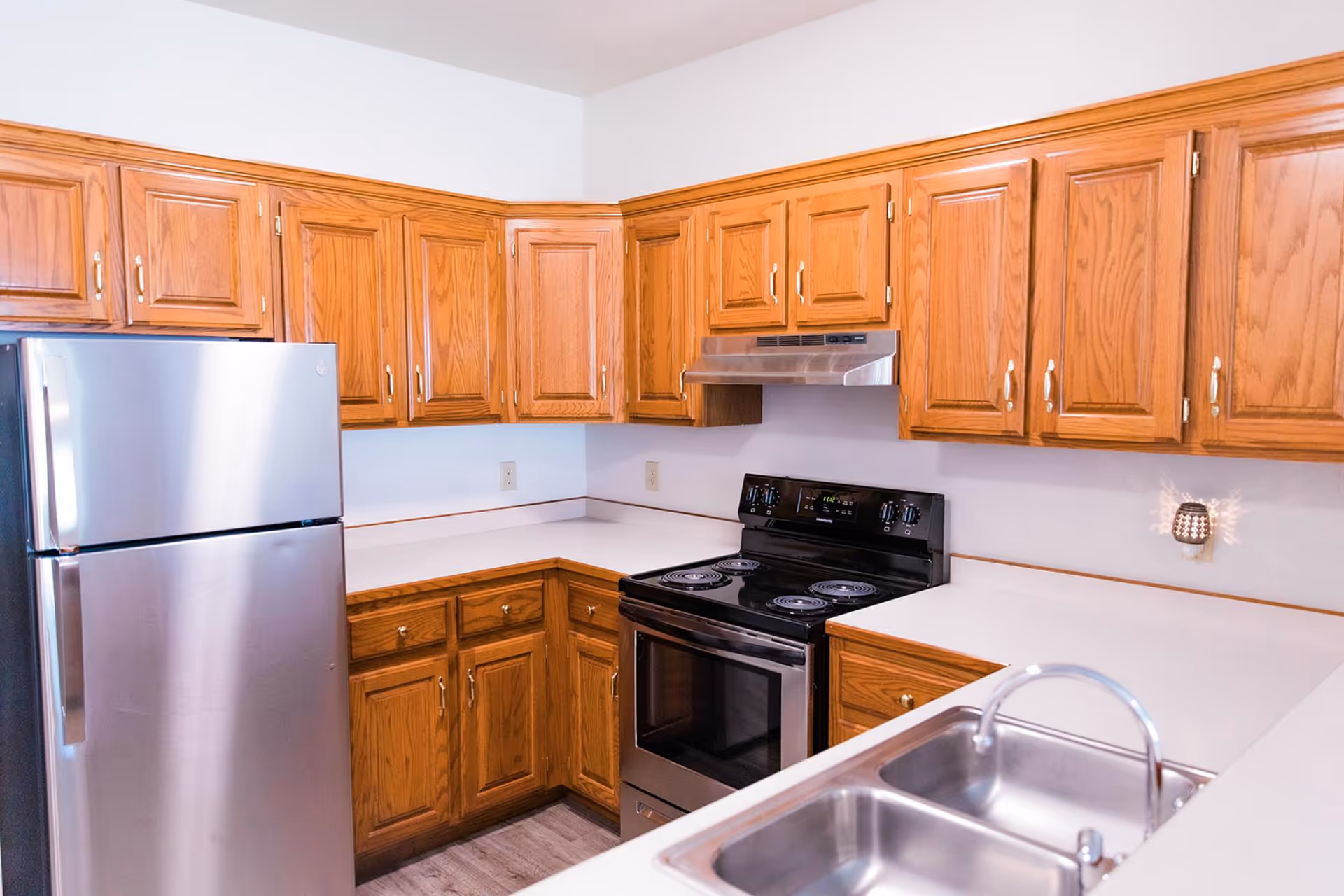 A kitchen with wooden cabinets, a stainless steel refrigerator, a black electric stove with an overhead stainless steel range hood, white countertops, and a double stainless steel sink with a curved faucet.