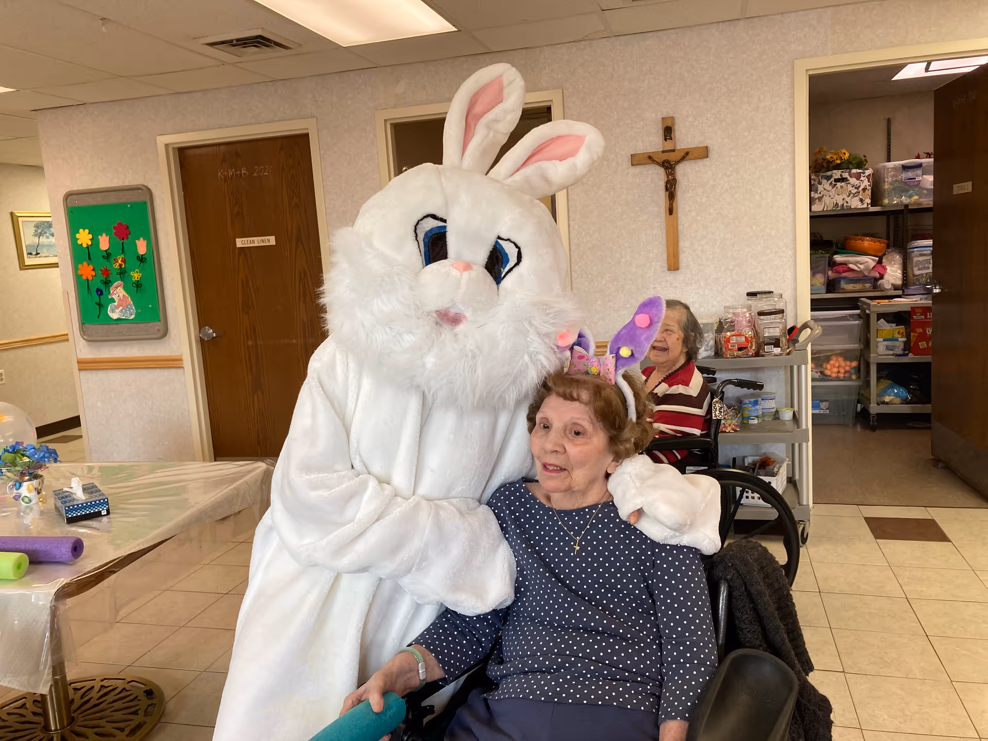 An elderly woman in a wheelchair wearing purple bunny ears is being hugged by a person dressed in a large white Easter Bunny costume inside a senior living facility. Another elderly woman is seated in a wheelchair in the background near a cart with various items. The room has tiled floors, a crucifix on the wall, and a door labeled 'Clean Linen'.