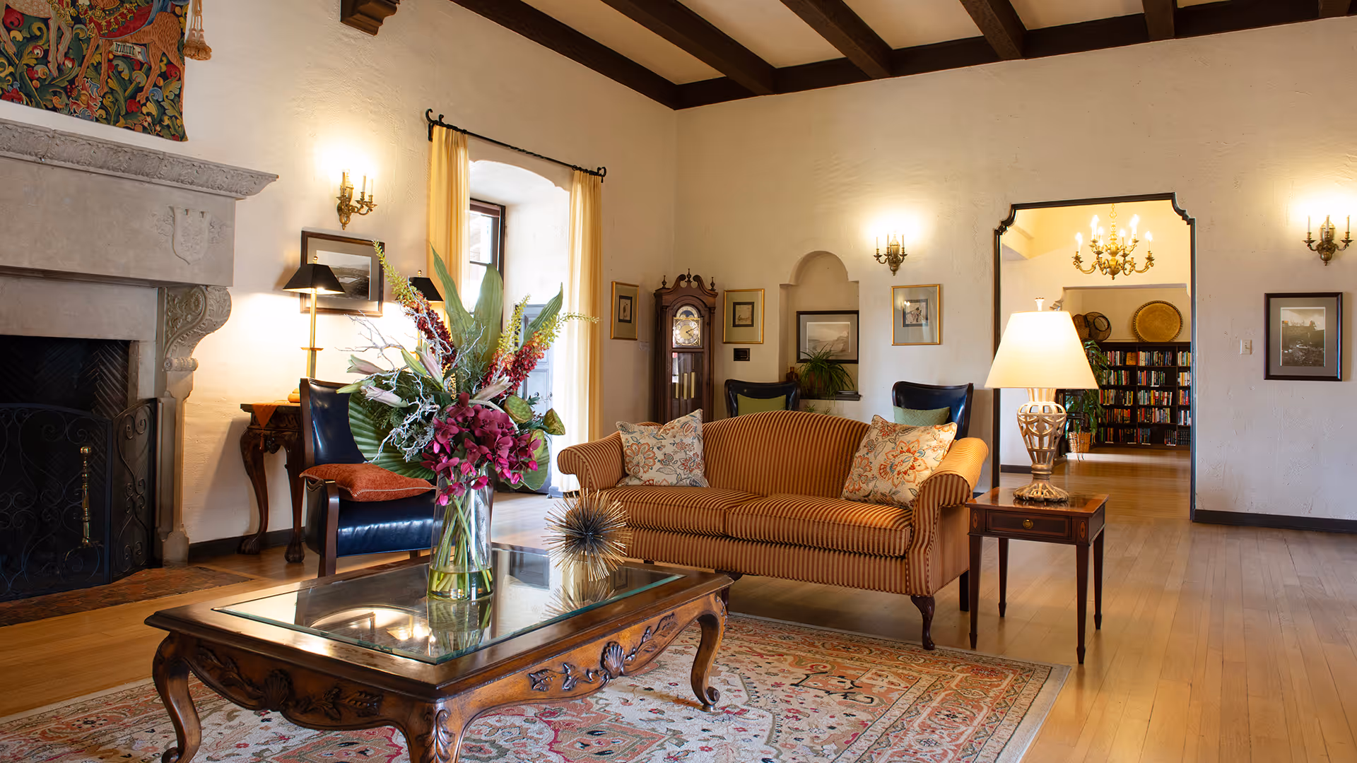 A cozy living room with a striped sofa adorned with patterned pillows, a wooden coffee table with a glass top holding a vase of colorful flowers, two dark leather chairs, a grandfather clock, and a fireplace with a decorative mantle. The room features wooden beams on the ceiling, wall sconces, framed pictures, and an open doorway leading to a room with bookshelves and a chandelier.