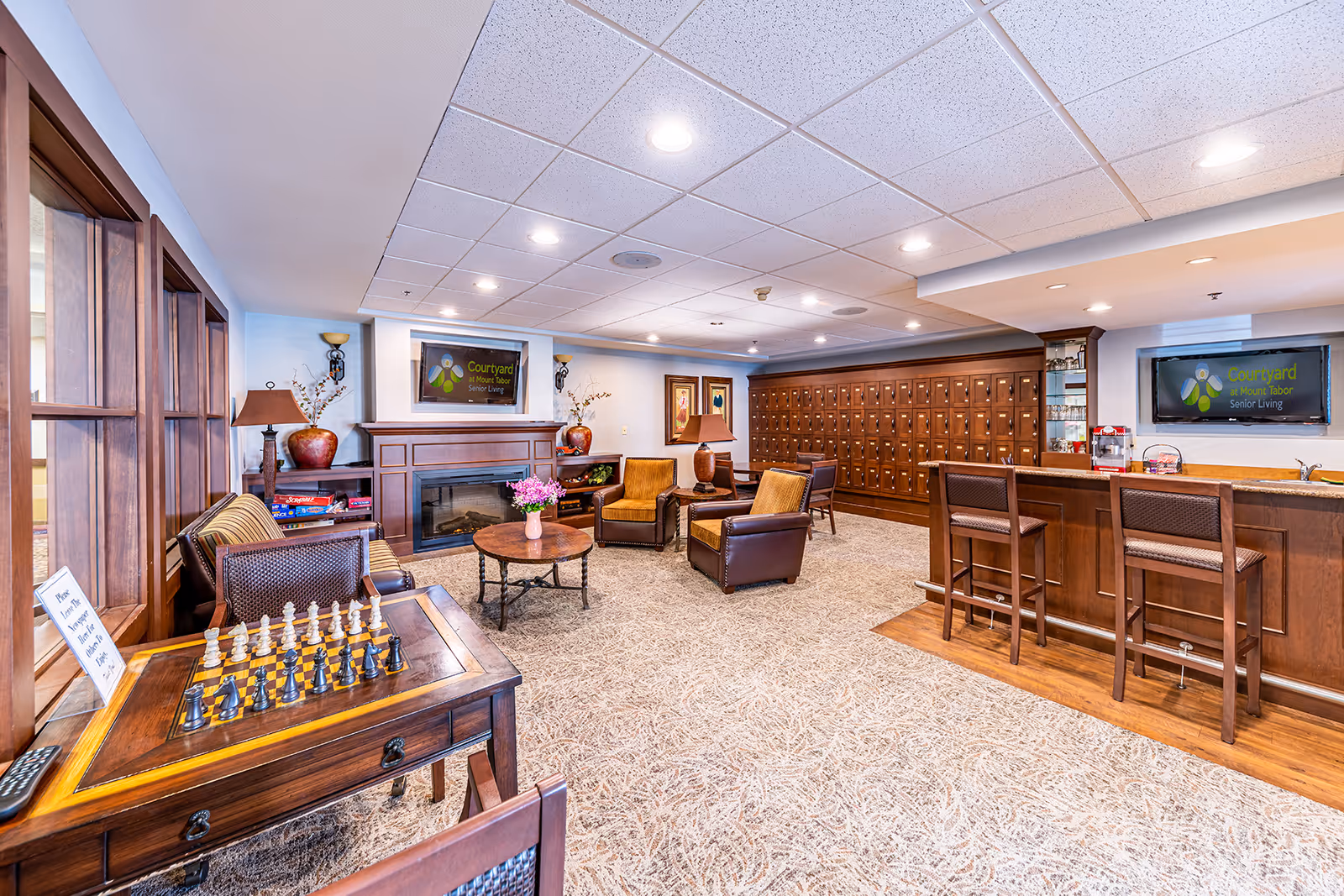 A well-appointed senior living lounge with armchairs around a fireplace, a chess table, and a bar counter with mail cubbies along the back wall.