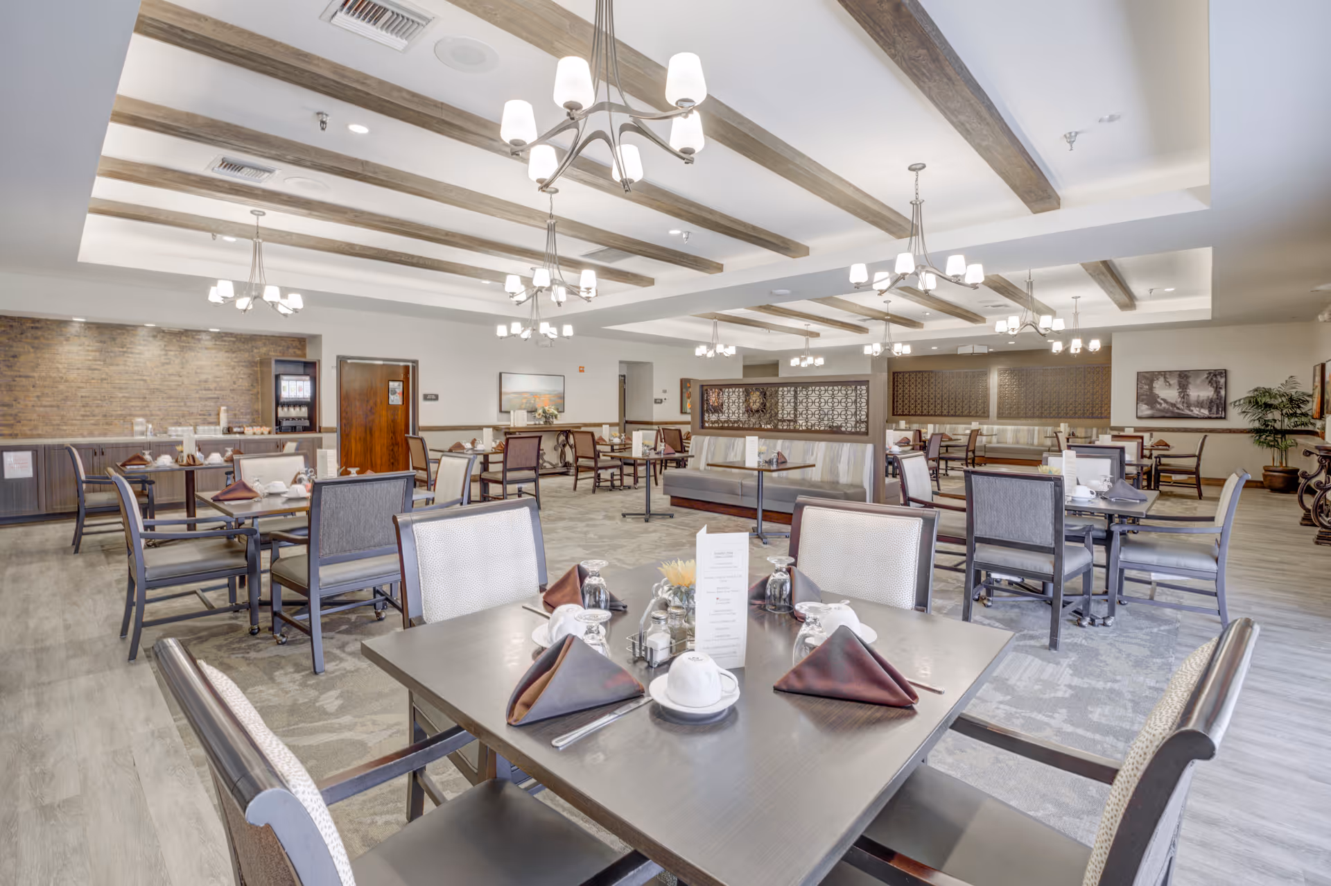 A spacious and well-lit dining room in an assisted living facility with multiple tables set with cups, napkins, and utensils. The room features wooden ceiling beams, modern chandeliers, and a mix of chairs and booth seating. The walls have decorative elements and artwork, and the floor is covered with a combination of carpet and wood-like flooring.