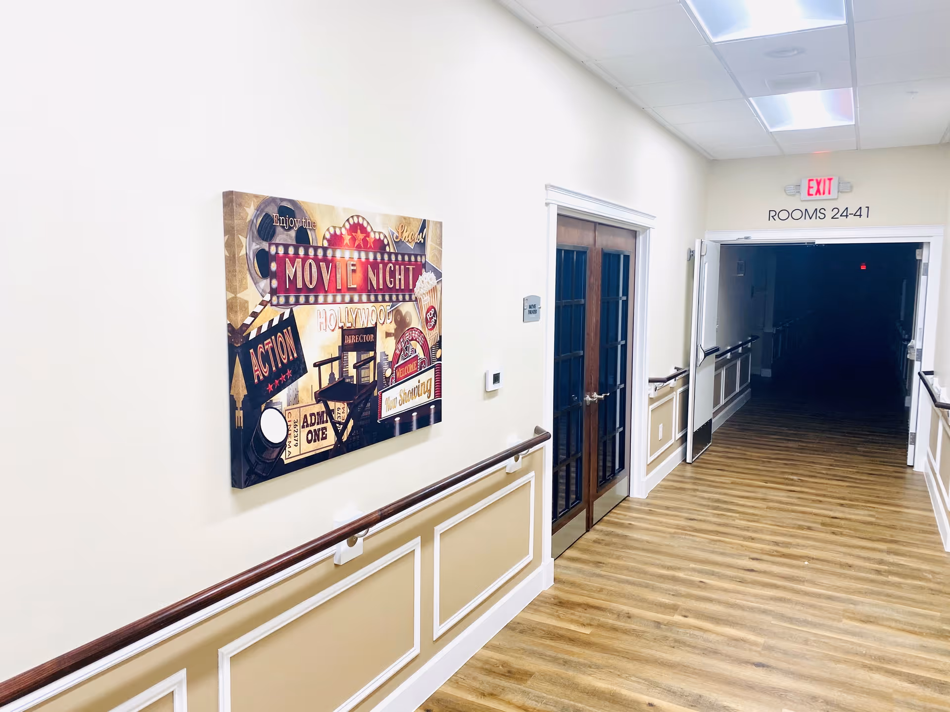 A well-lit hallway in a senior living facility with wooden flooring and beige walls with white trim. On the left wall, there is a colorful movie night-themed poster. At the end of the hallway, double doors with glass panels are visible, and an exit sign above a doorway indicates rooms 24-41. Handrails run along both sides of the hallway.