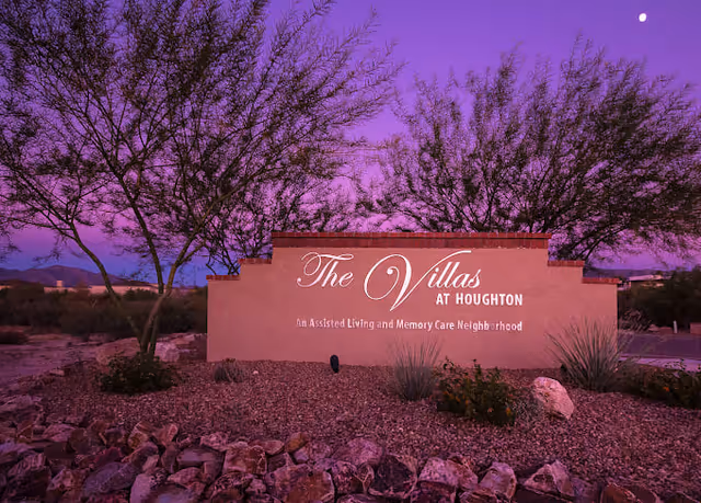 Entrance sign for The Villas at Houghton, an assisted living and memory care neighborhood, surrounded by desert landscaping with rocks and trees under a purple twilight sky.