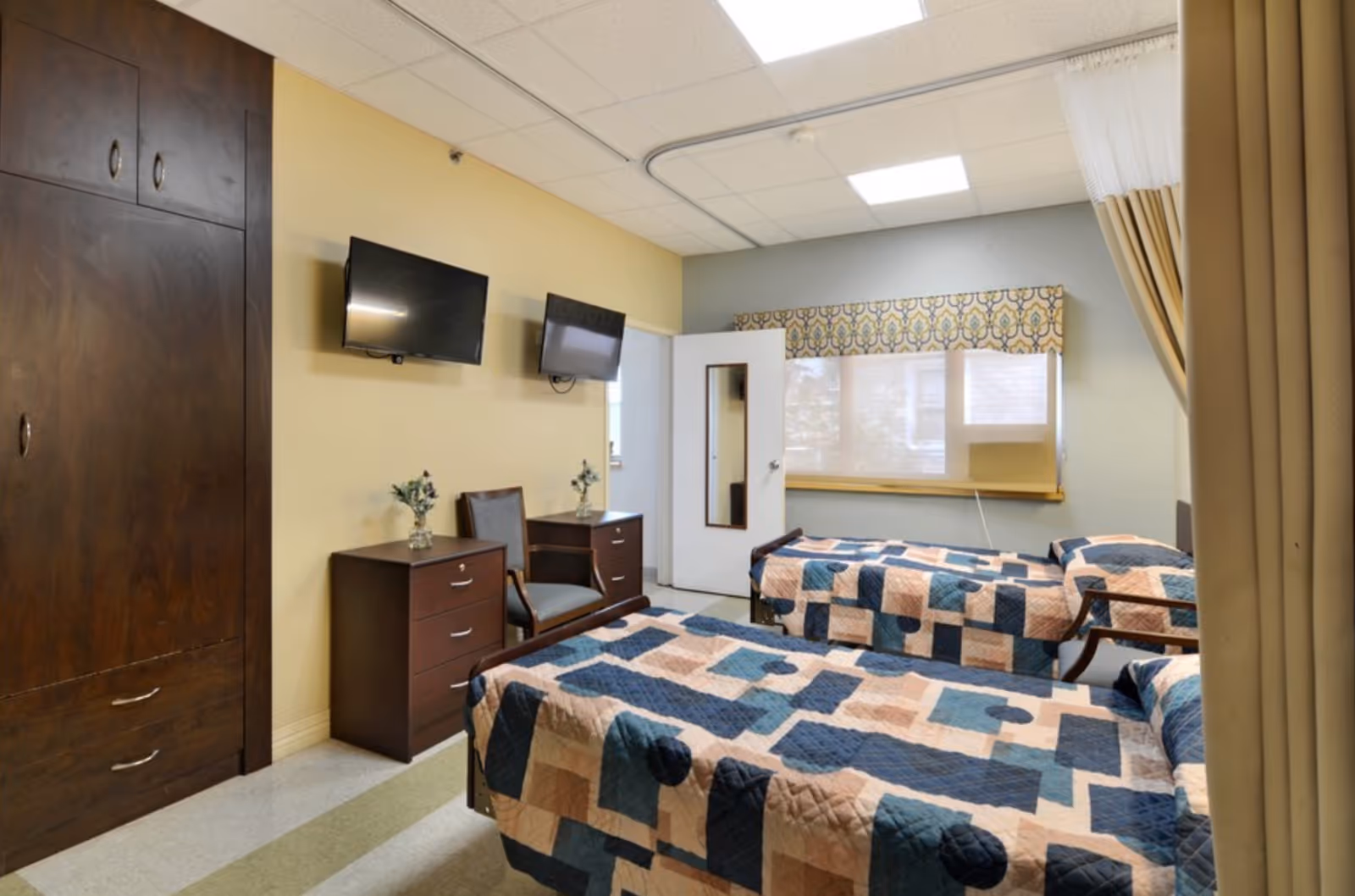 A senior living facility bedroom with two single beds covered in patterned quilts in shades of blue, beige, and peach. The room has a window with a decorative valance, two wall-mounted flat-screen TVs, a wooden chair, two small wooden dressers with vases of flowers, and a large wooden wardrobe. The ceiling has recessed lighting, and a privacy curtain is partially visible on the right side.