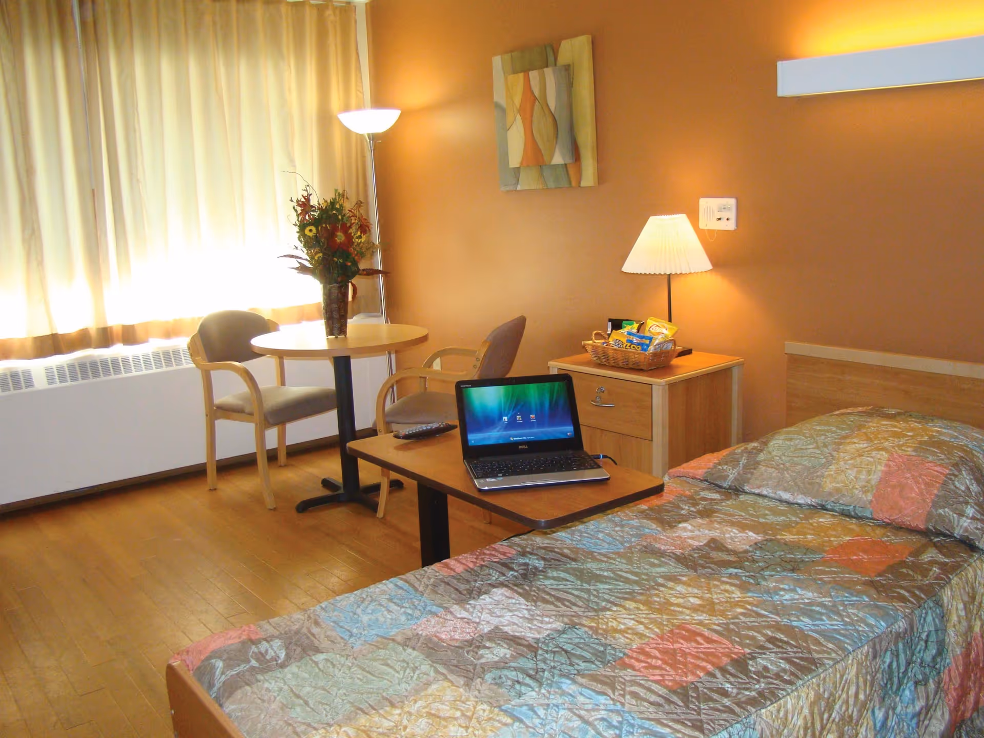 A cozy bedroom in a senior living facility with a single bed covered in a colorful quilt. Next to the bed is a wooden nightstand with a lamp and a basket of snacks. A small table with a laptop and remote control is positioned at the foot of the bed. In the background, there is a round table with two chairs and a vase of flowers, illuminated by a floor lamp near a window with beige curtains.