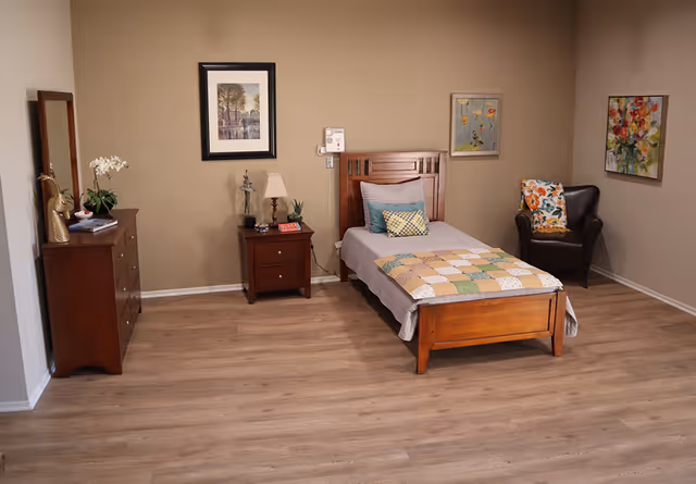 Single-occupancy assisted living bedroom with a wooden twin bed, nightstand, dresser, and armchair against beige walls.