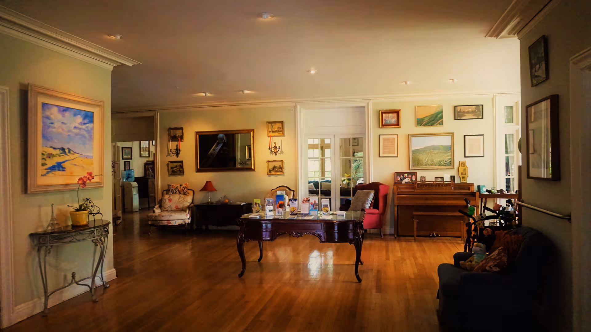 A warmly lit interior room with wooden flooring, featuring a central wooden table displaying brochures. The room is decorated with various framed paintings and pictures on the walls. There are several chairs, including a floral armchair, a red armchair, and a blue sofa with stuffed toys. A piano is positioned against the far wall, and a small decorative table with a potted plant is on the left side.