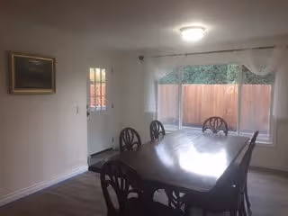 A dining room with a wooden dining table and six matching chairs. There is a large window with sheer white curtains overlooking a fenced backyard. A door with glass panels is visible on the left wall, and a framed painting hangs on the adjacent wall. The room has light-colored walls and wood flooring.