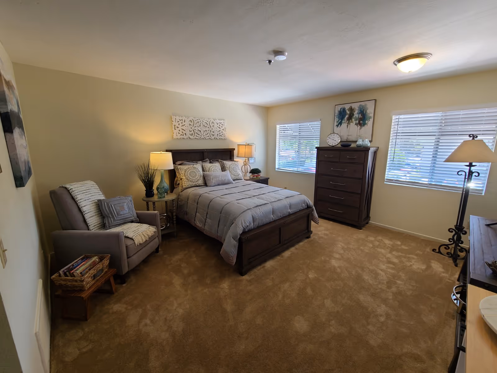 A cozy bedroom with a queen-sized bed featuring multiple pillows and a gray comforter. There is a cushioned armchair with a throw blanket and pillow on the left side, next to a round side table with a lamp and decorative plant. On the right side of the room, there is a tall dark wooden dresser with decorative items on top, two windows with blinds letting in natural light, and a floor lamp. The walls are painted light beige and the floor is carpeted in a light brown color.