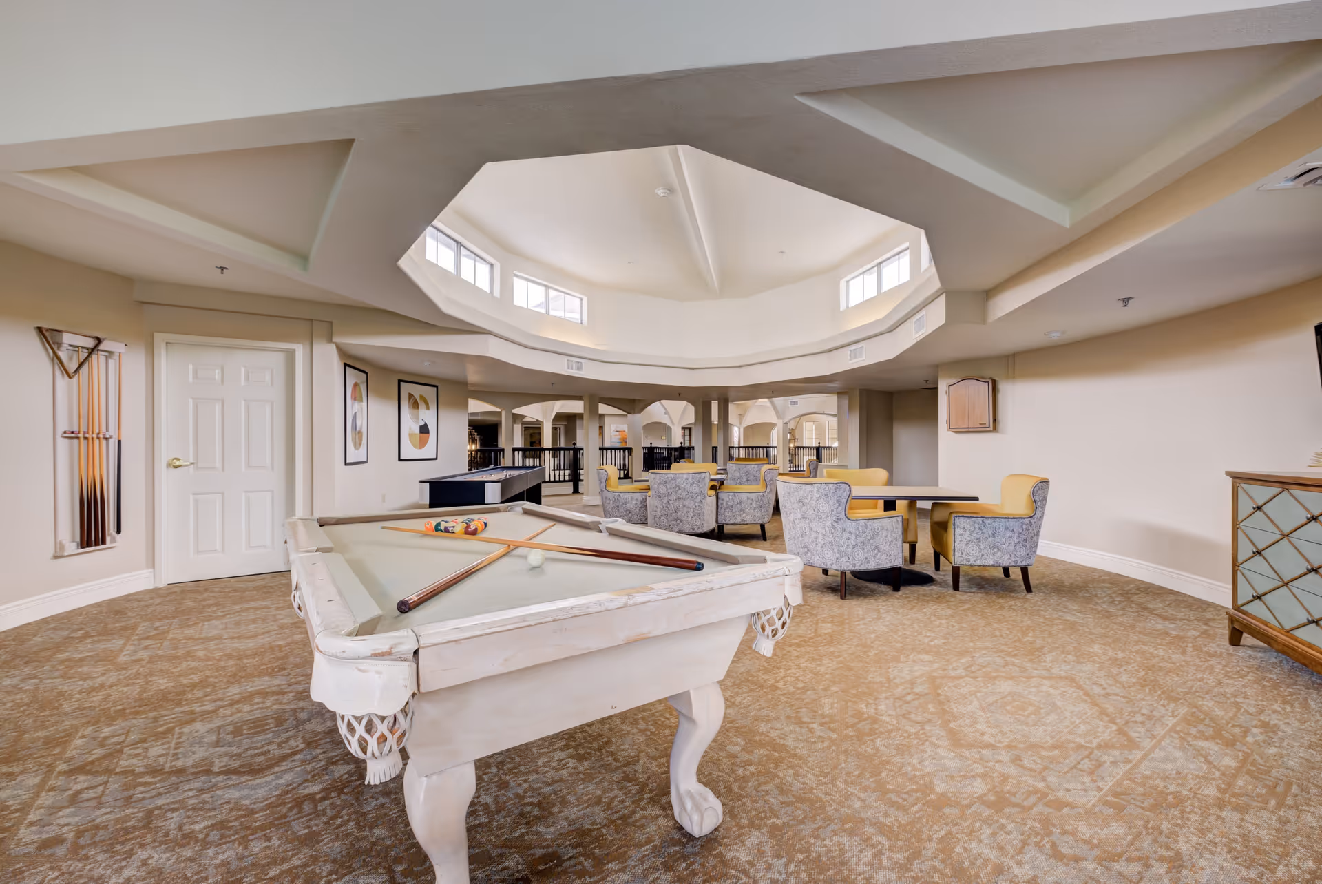 A spacious recreational room with a white pool table in the foreground, pool cues mounted on the wall, and a seating area with tables and upholstered chairs. The room features a unique octagonal ceiling with windows allowing natural light to enter.
