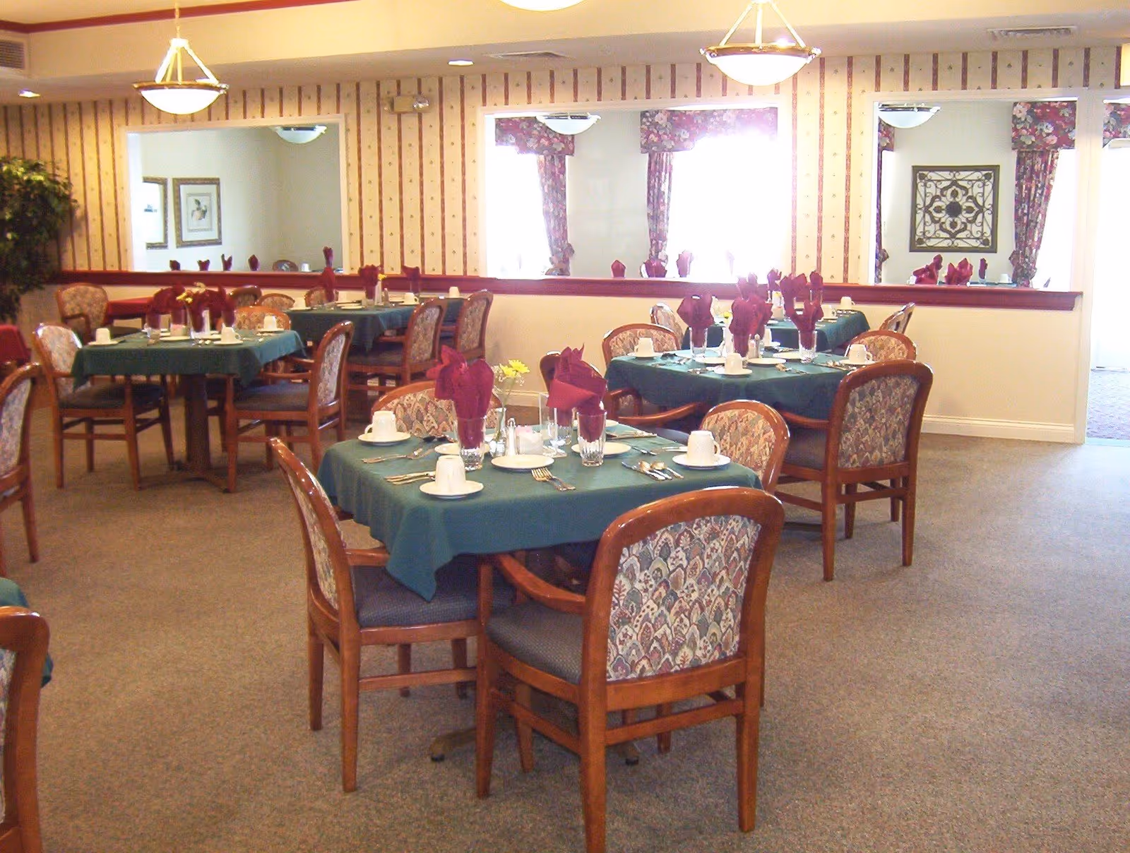 Dining room with multiple round tables set with green tablecloths, folded napkins, cups, and wooden chairs.