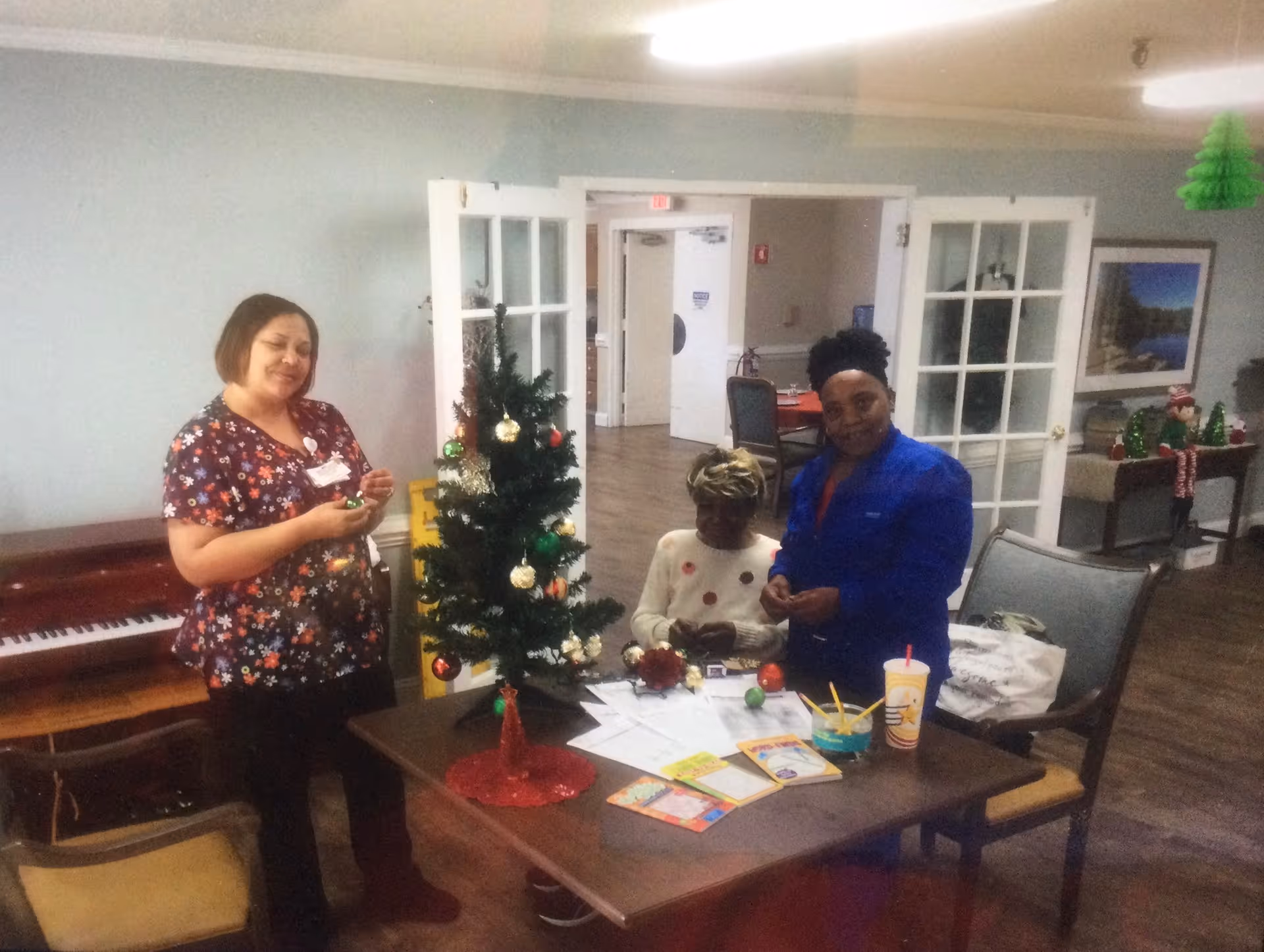 Three women decorating a small Christmas tree on a table in a room with wooden flooring. One woman is standing on the left wearing a floral top, another woman is seated at the table, and the third woman is standing on the right wearing a blue jacket. The table has papers, cards, and a drink cup on it. In the background, there is a piano, chairs, and a table with holiday decorations.