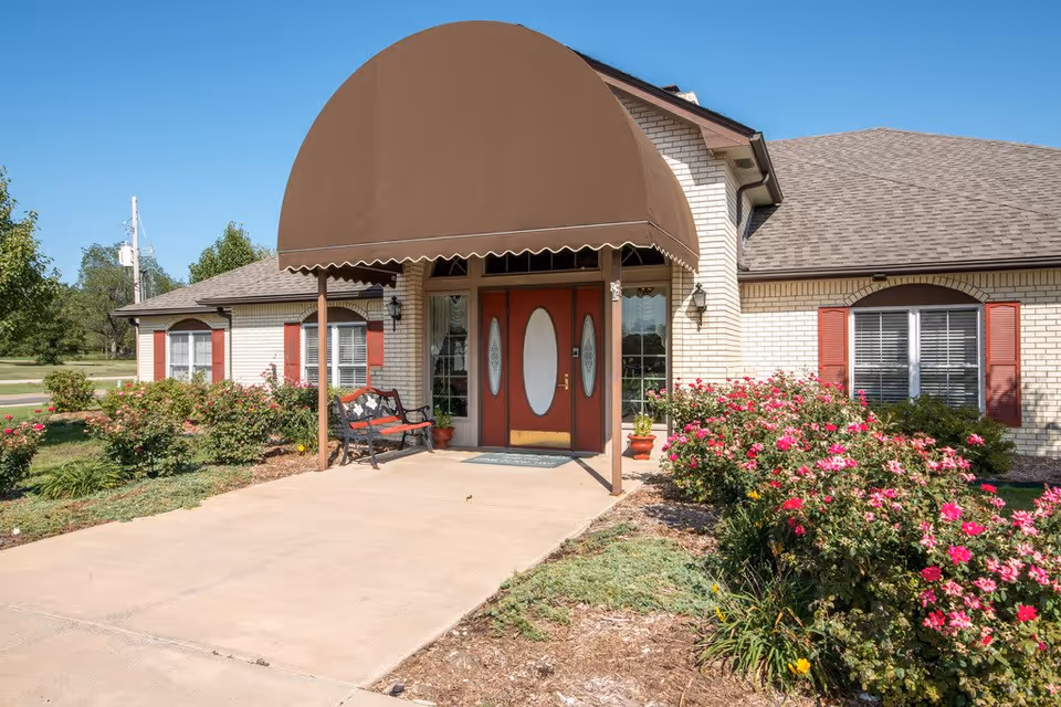 Front entrance of a senior living facility with a brown awning over double red doors with oval glass panels. There is a bench to the left of the entrance and flower beds with pink flowers on either side of the walkway leading to the door. The building has light-colored brick walls and red shutters on the windows.