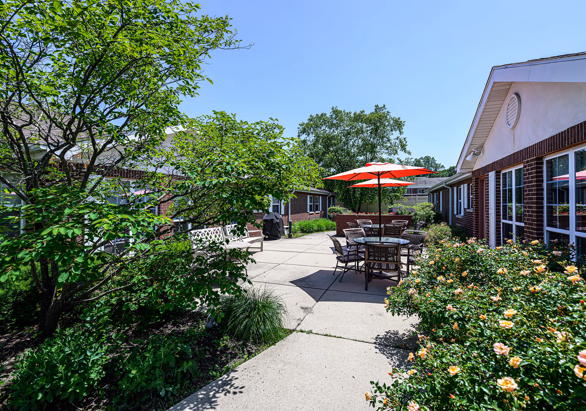 Outdoor patio area at Arden Courts - ProMedica Memory Care Community (Old Orchard) featuring a concrete walkway, green trees and bushes, a round table with chairs under a red umbrella, and brick buildings on either side under a clear blue sky.