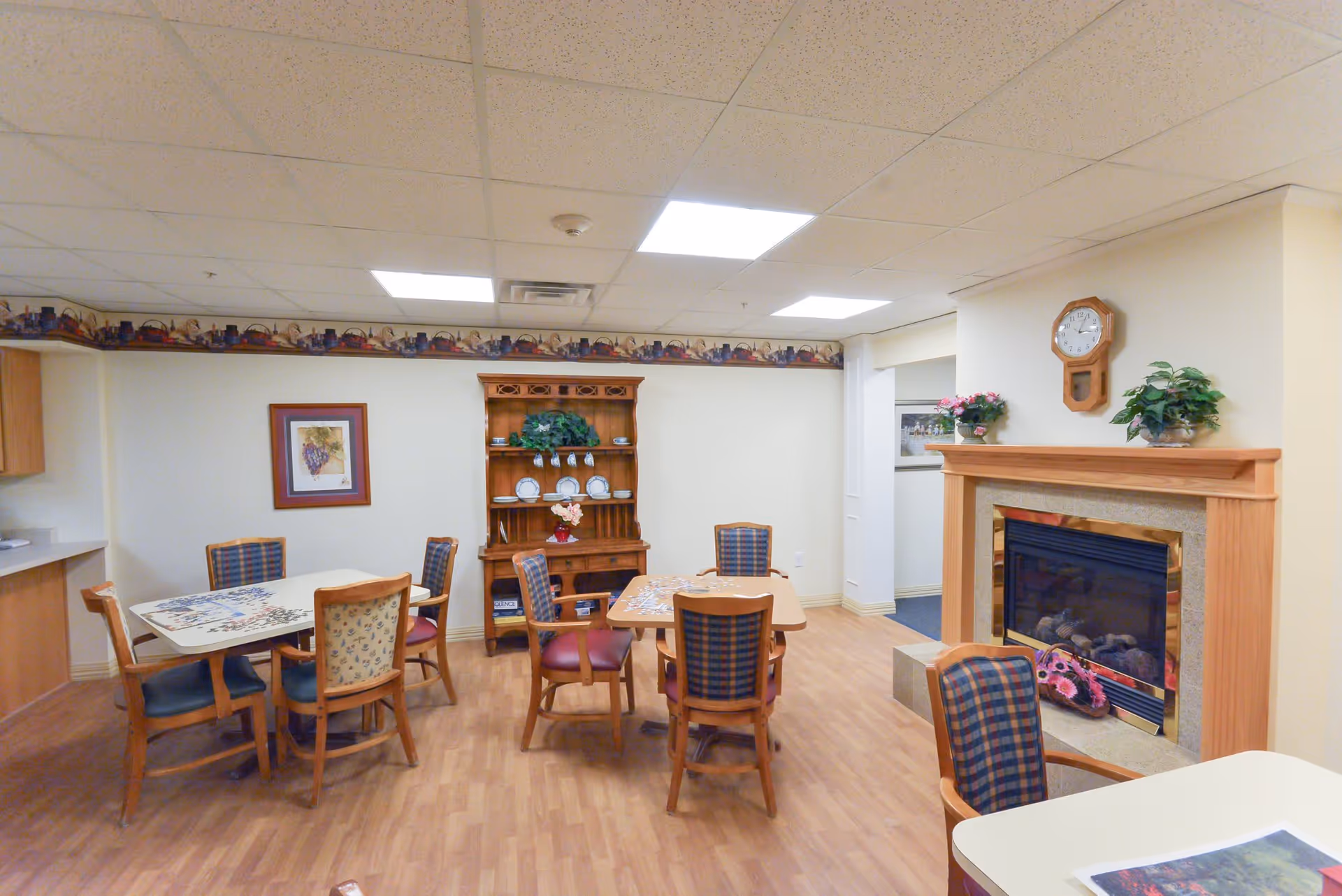 A cozy common area in an assisted living facility featuring two tables with chairs around them, a wooden hutch displaying plates and cups, a fireplace with a wooden mantle decorated with plants and a clock, and a framed picture on the wall. The room has wood flooring and a drop ceiling with fluorescent lighting.