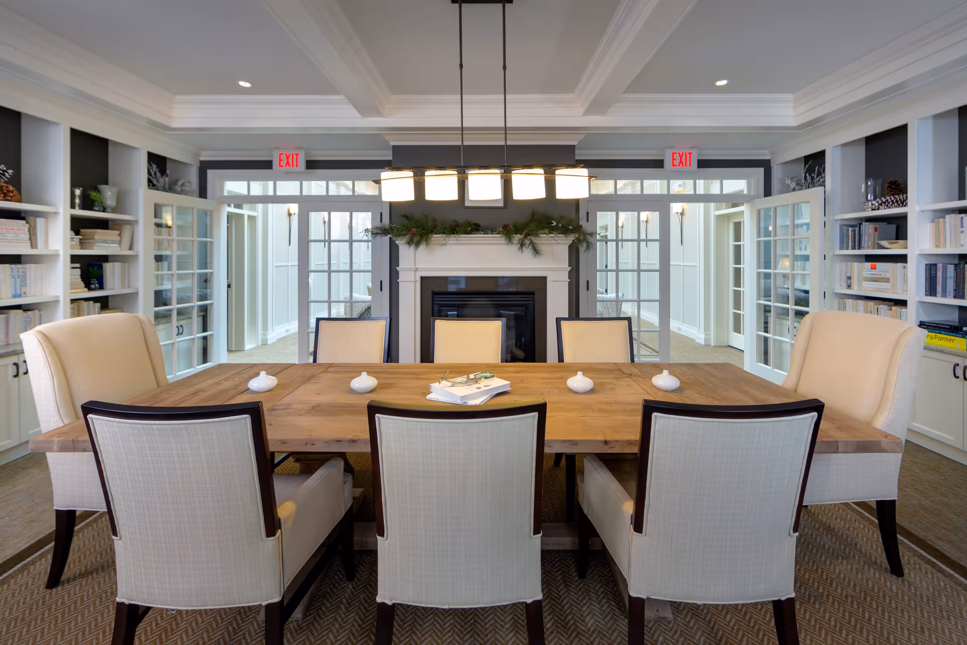 A well-lit dining room with a large wooden table surrounded by eight upholstered chairs. The room features built-in white bookshelves filled with books and decorative items on both sides. At the far end, there is a fireplace decorated with greenery and a modern light fixture hanging above the table. Glass doors and windows allow natural light to enter, and exit signs are visible above the doors.