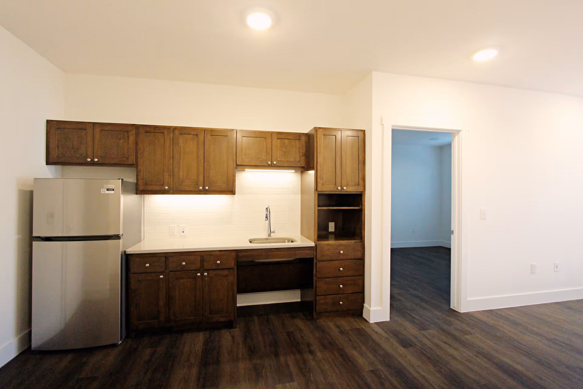 Small kitchenette with a stainless steel refrigerator, wooden cabinets, countertop sink, and dark wood flooring in an open bright room.