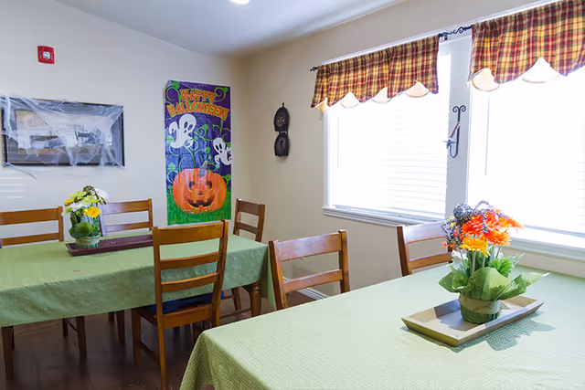 A dining room with two tables covered in green tablecloths, each with a floral centerpiece. There are wooden chairs around the tables. The room has two windows with plaid valances letting in natural light. Halloween decorations, including a colorful 'Happy Halloween' poster with ghosts and a pumpkin, are displayed on the wall.